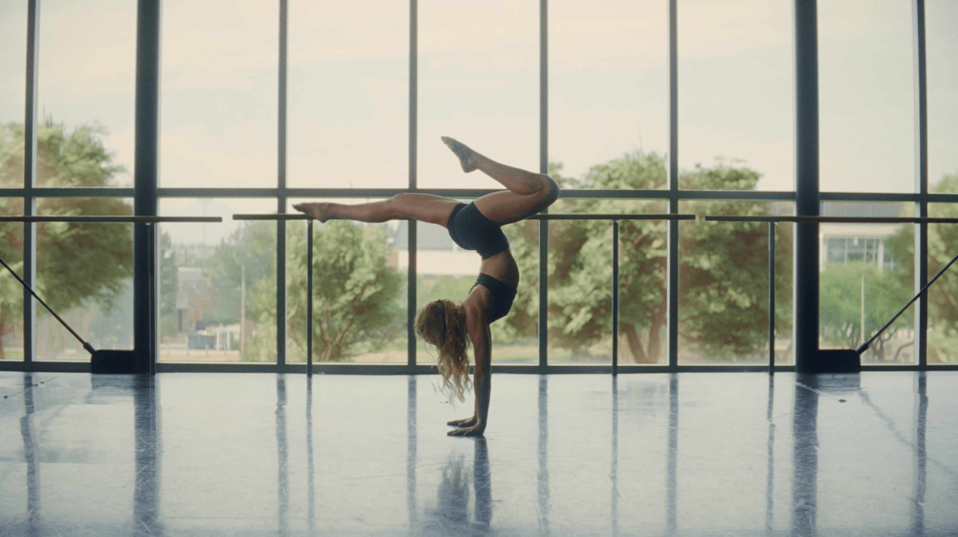 A dancer in a handstand at the barre of a sunlit studio, floor-to-ceiling windows onto Phoenix greenery behind her