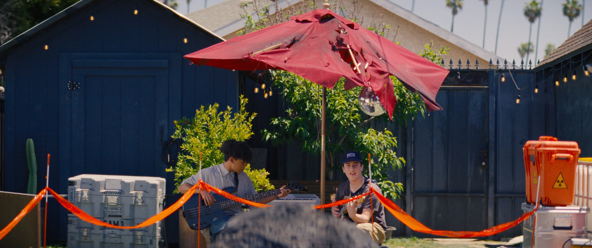 Two teenagers playing bass and keys under a red patio umbrella in a suburban backyard, from the short film Dog Sees God
