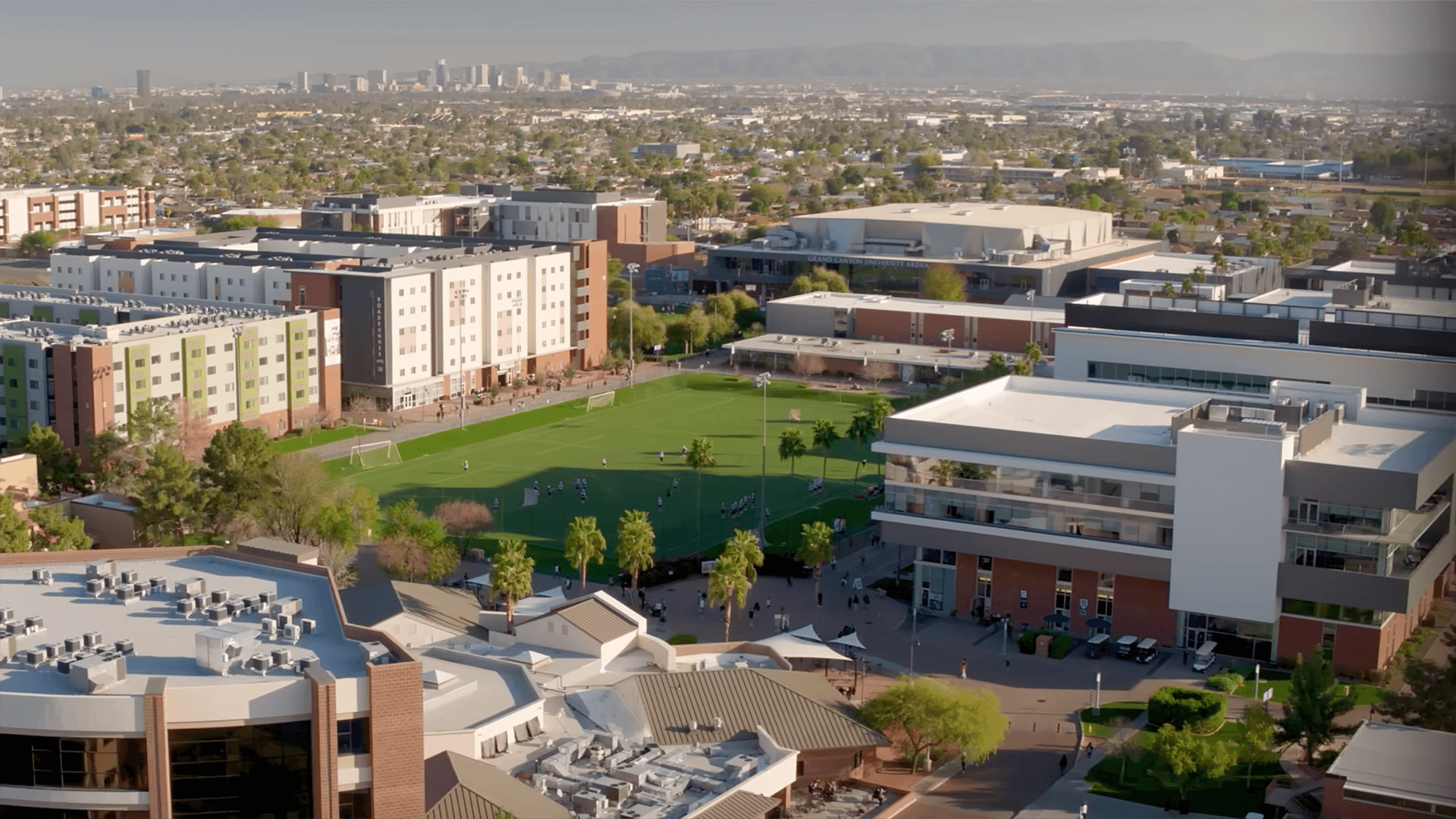 Aerial over the Grand Canyon University campus in Phoenix — soccer pitch, residence halls, and the downtown skyline beyond