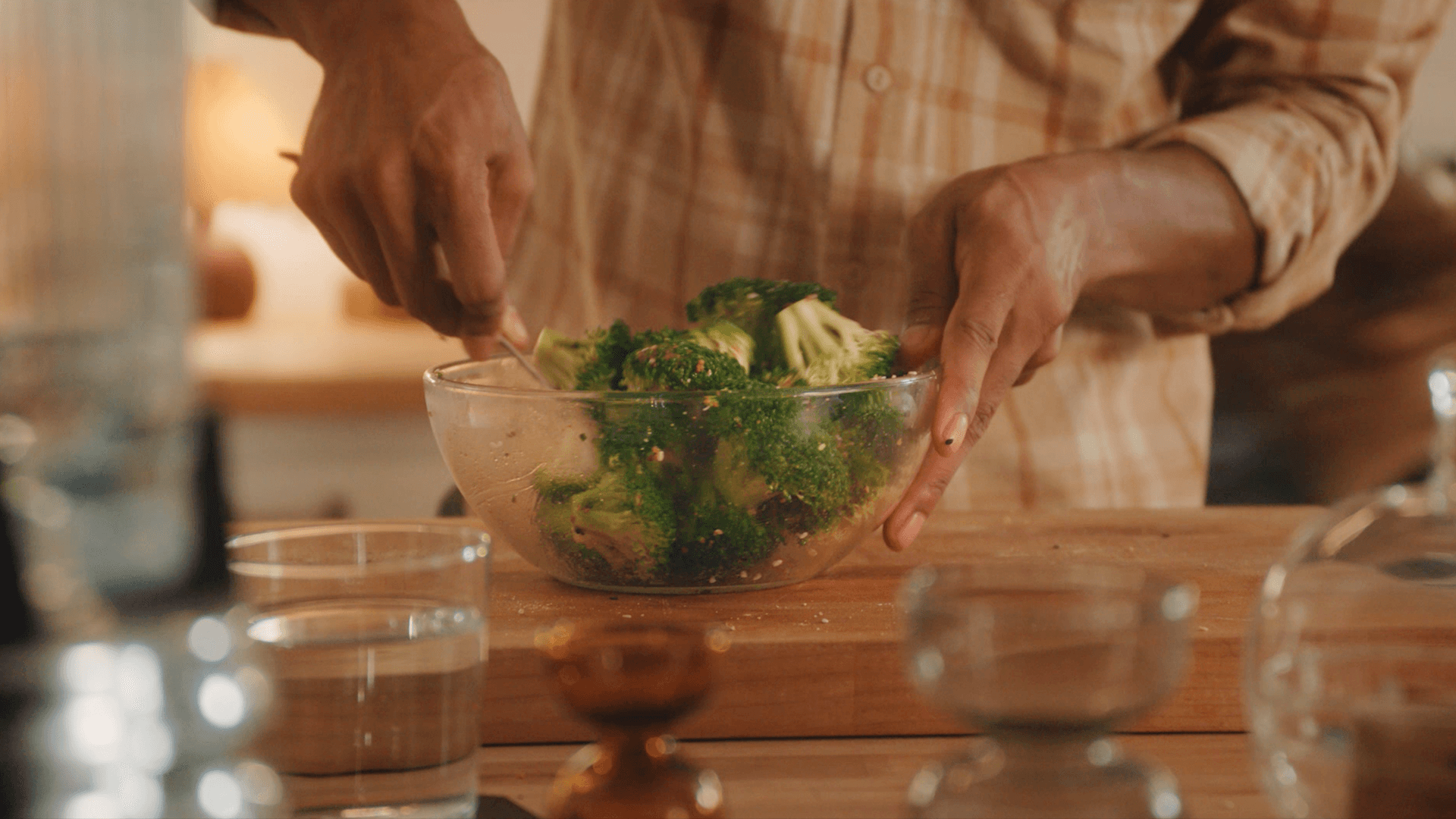 Hands lifting a glass bowl of roasted, seasoned broccoli off a wooden kitchen counter in warm evening light