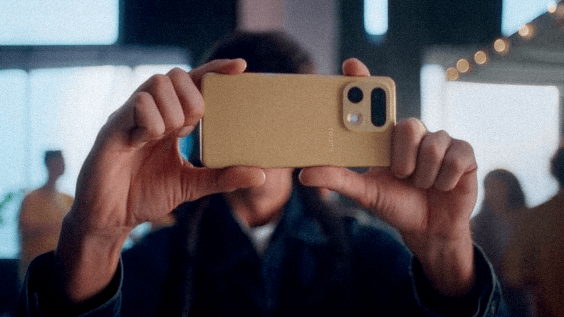 Hands holding a gold Realme 16 Pro sideways to film, a warm bar interior soft behind it