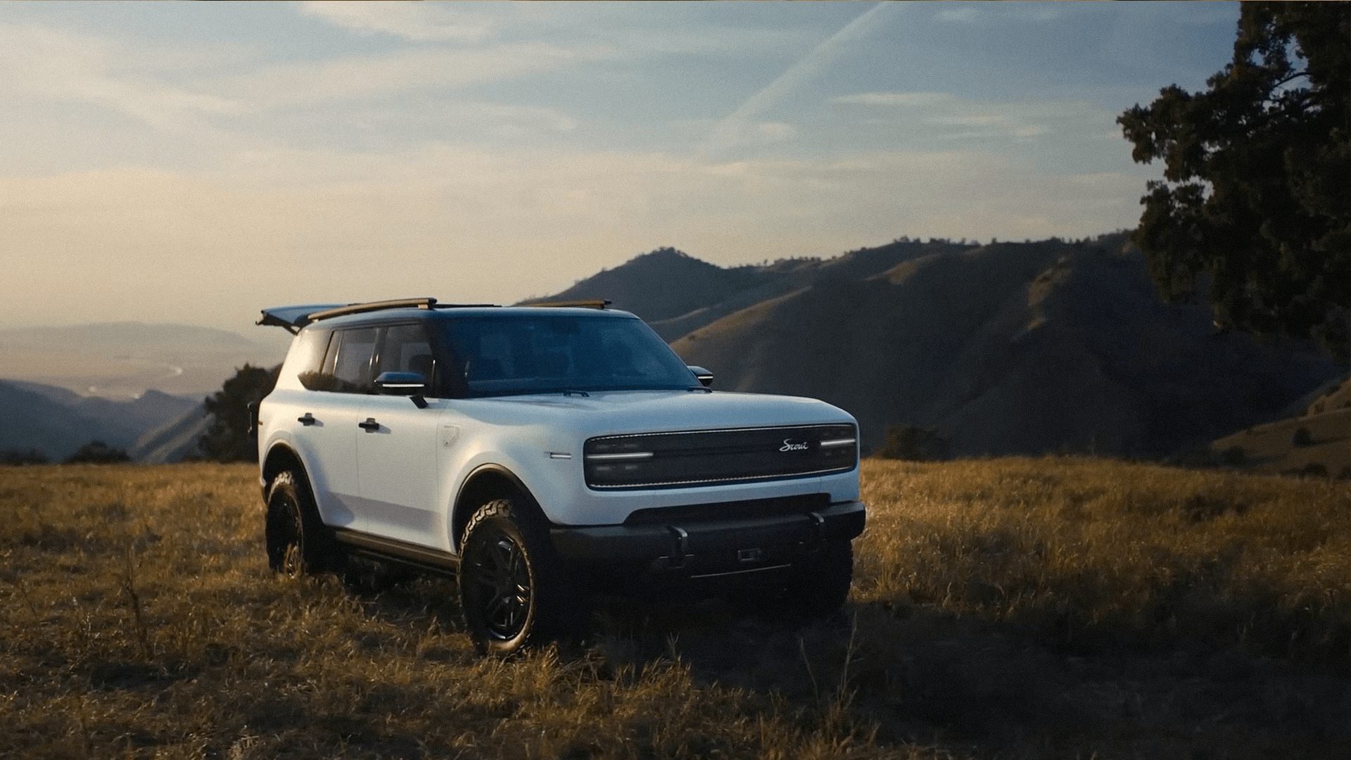 A white Scout Traveler parked in dry golden grass on a ridge, the oak hills of Tejon Ranch falling away behind it at dusk