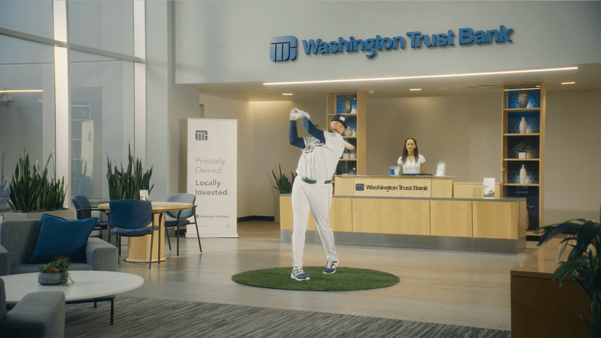 Cal Raleigh, in a Washington Trust Bank uniform, taking a full swing off a patch of turf in the middle of a bank lobby